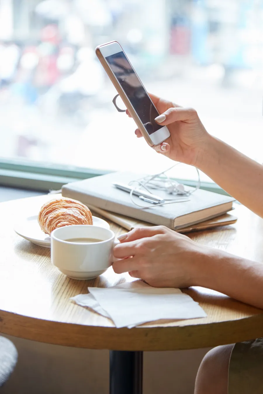 A guest holding a phone at a café table beside a coffee and a croissant